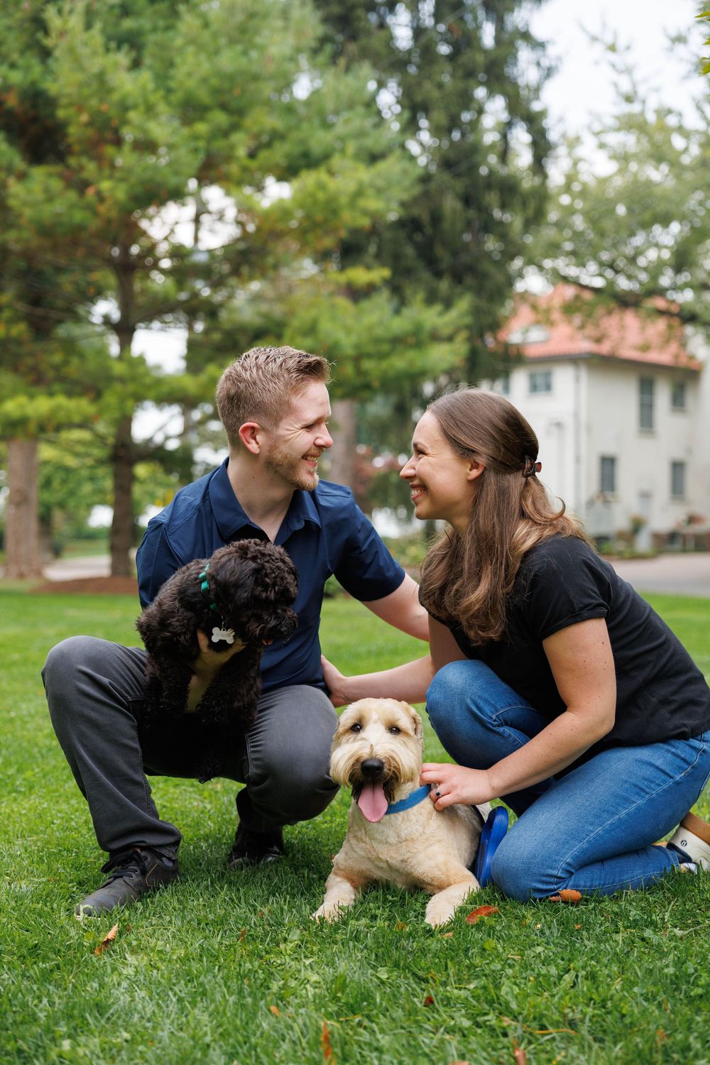 Couple sits on grass with their two dogs, a black dog and a light-colored terrier, enjoying a sunny day outdoors.