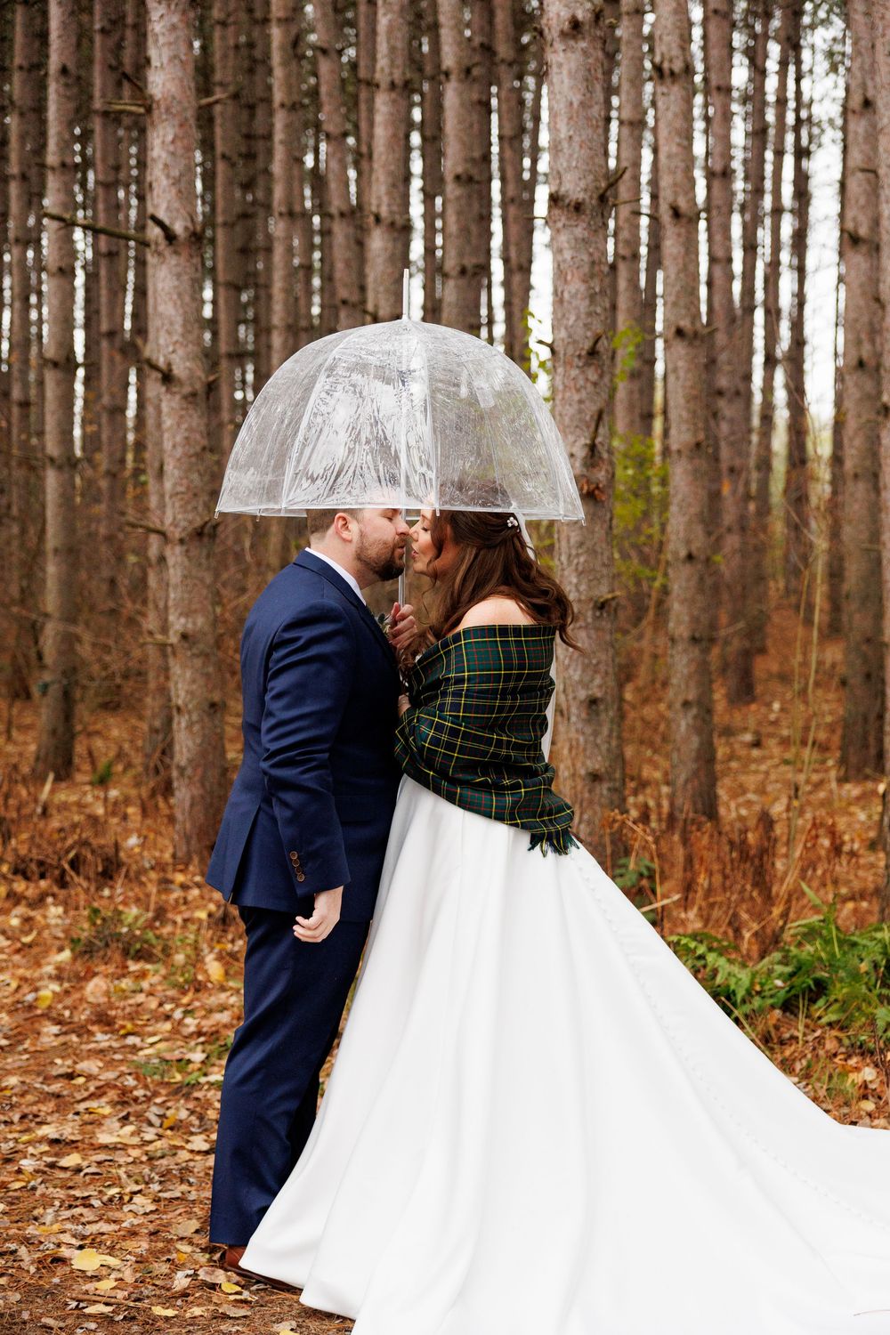 A romantic wedding photo series in a pine forest with fallen autumn leaves showing a couple in navy and white attire.