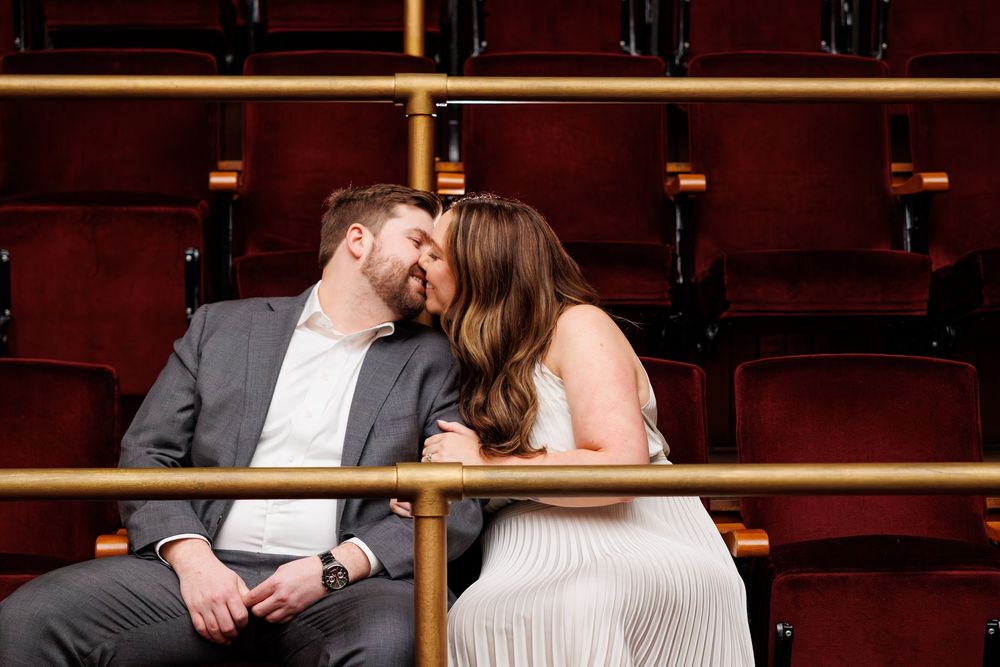 A couple shares a romantic moment in an elegant theater with rich red velvet seating and ornate decor.