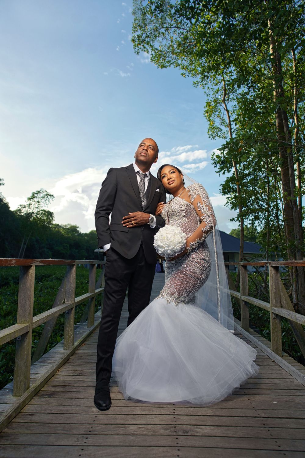 A romantic wedding couple poses together on a wooden bridge surrounded by lush green trees on their special day.