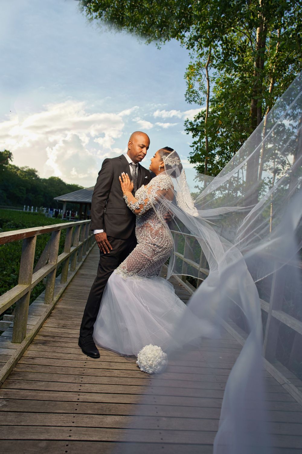 A romantic wedding couple shares an intimate moment on a wooden bridge with a flowing veil in the breeze.
