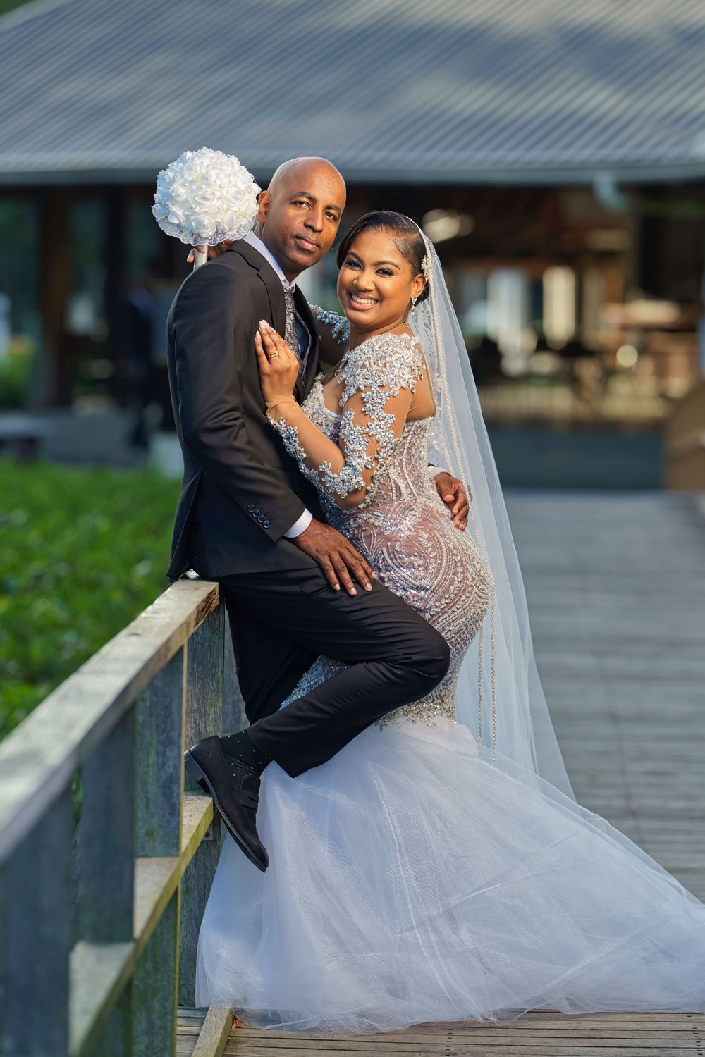 A bride in an elegant white lace gown and groom in a dark suit share romantic moments on a boardwalk.