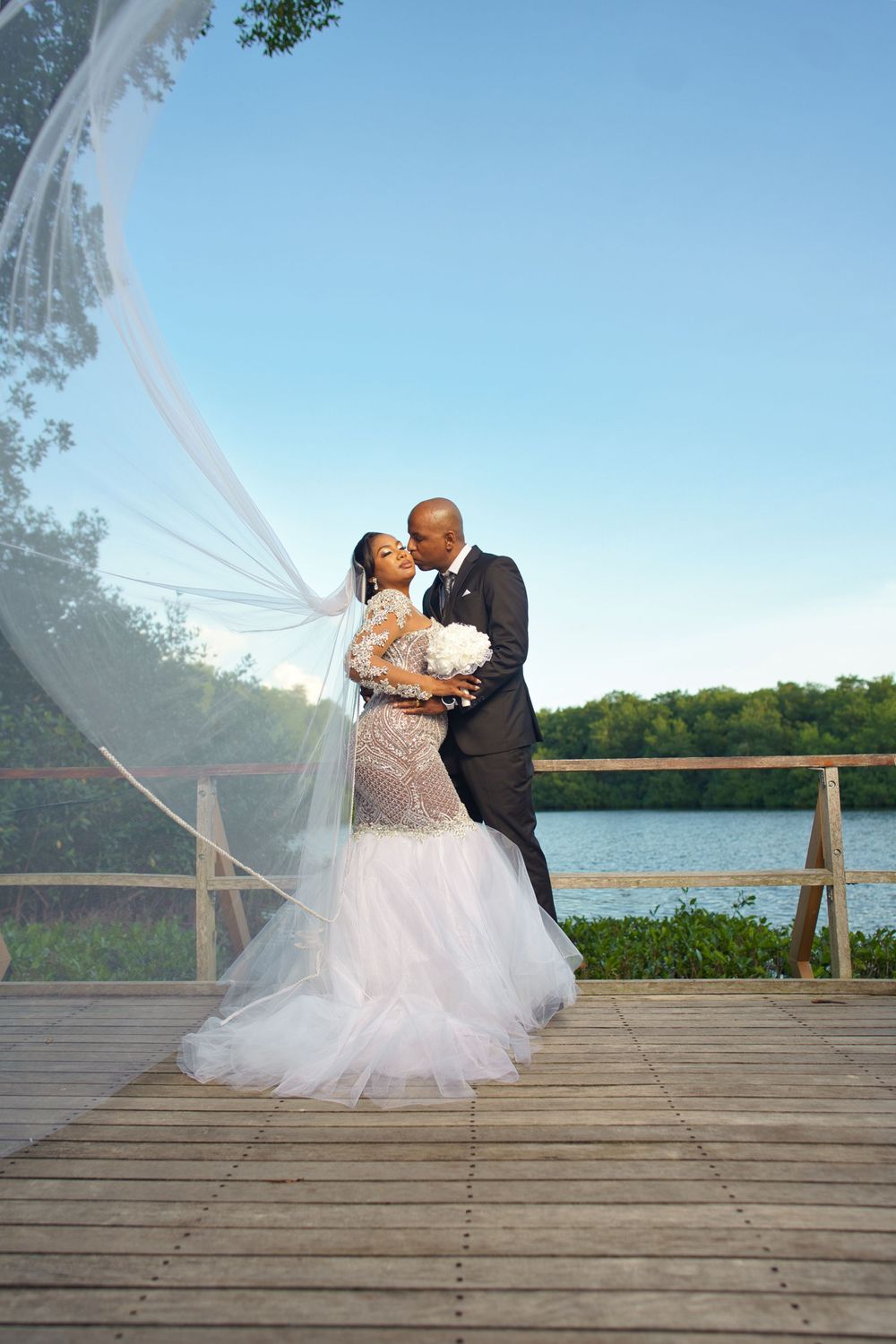 A romantic wedding photo of a couple embracing on a wooden dock by a lake with a flowing white veil in the breeze.