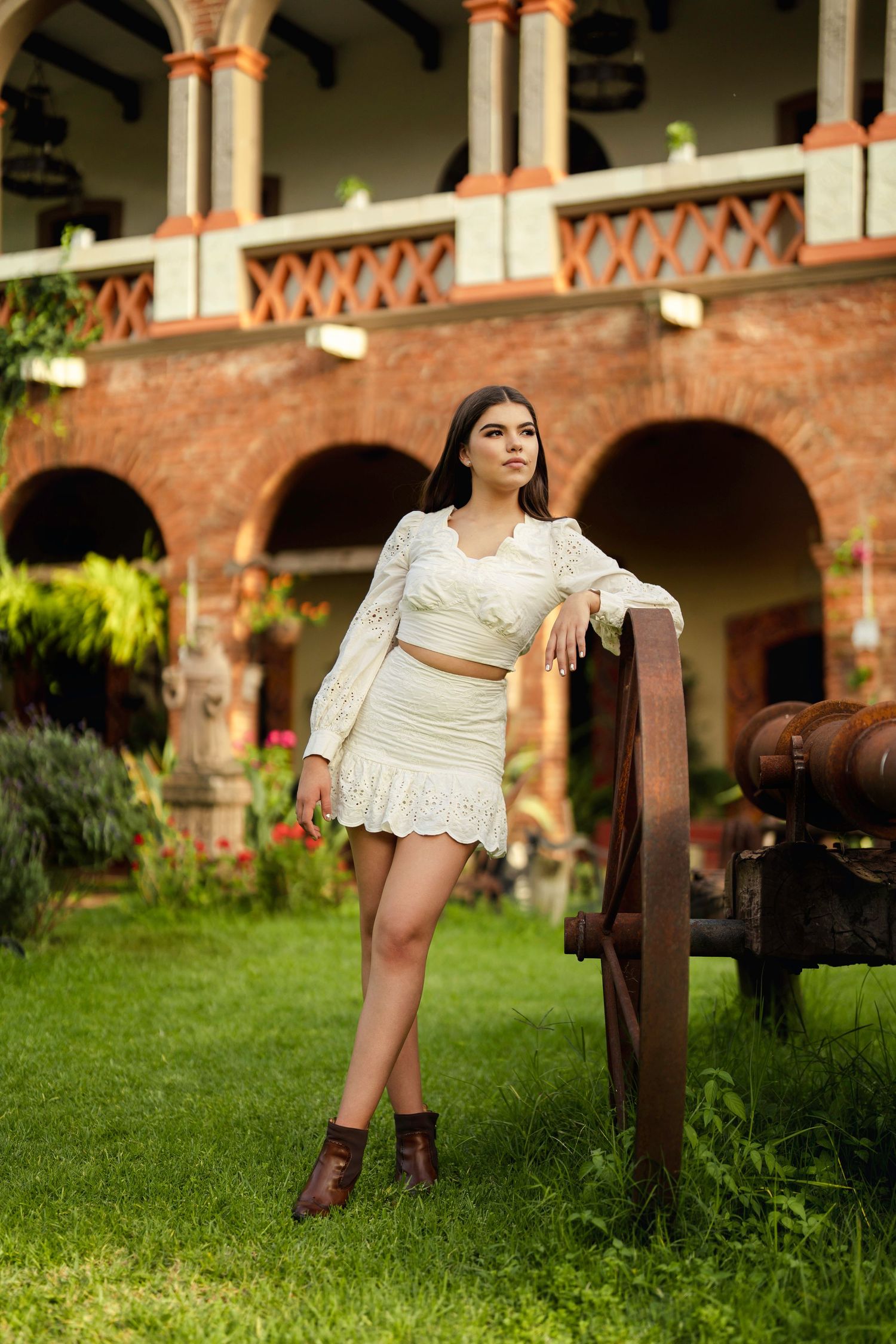 Una mujer vestida con un vestido blanco y botas posa en un jardín con elementos arquitectónicos rústicos al fondo