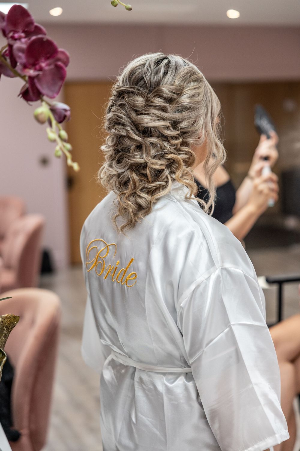 A white satin robe with 'Bride' embroidered in gold text is shown from behind with an elegant updo hairstyle.