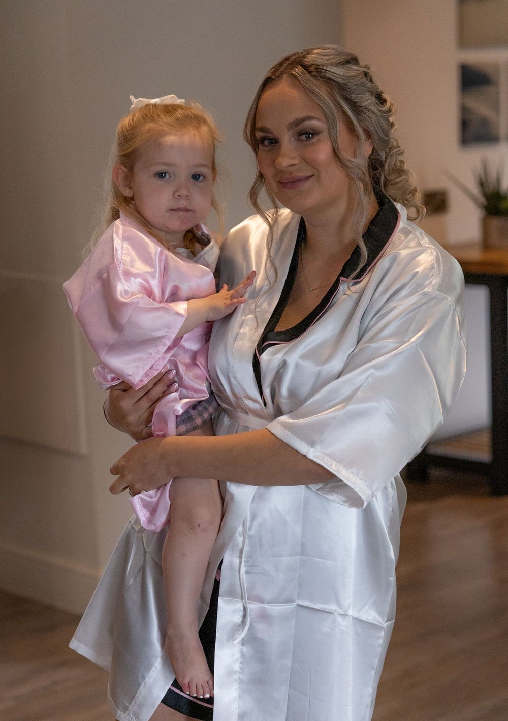 A woman in a white satin robe holds a small child in pink during wedding preparations.