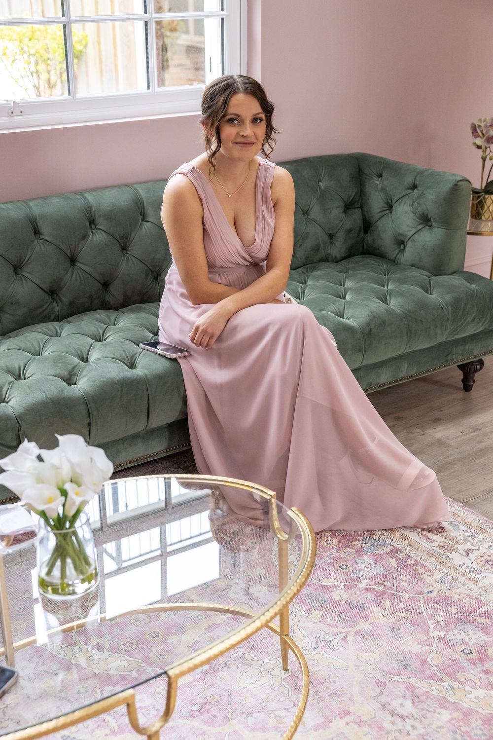 A pink dress-clad figure sits elegantly on a tufted green velvet sofa with gold coffee tables and white flowers nearby.