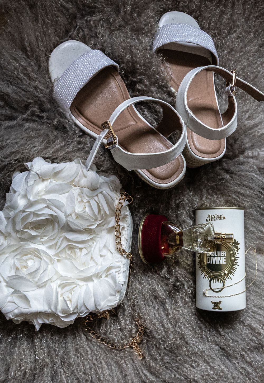 Wedding accessories including white sandals, a garter, and perfume bottles arranged on a textured gray surface.