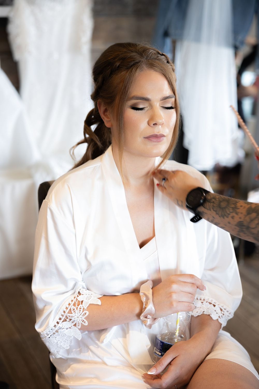 Bride in white lace robe getting her makeup professionally done during morning preparations.