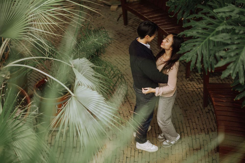 A romantic couple embraces in a lush greenhouse setting surrounded by tropical palm fronds and greenery.