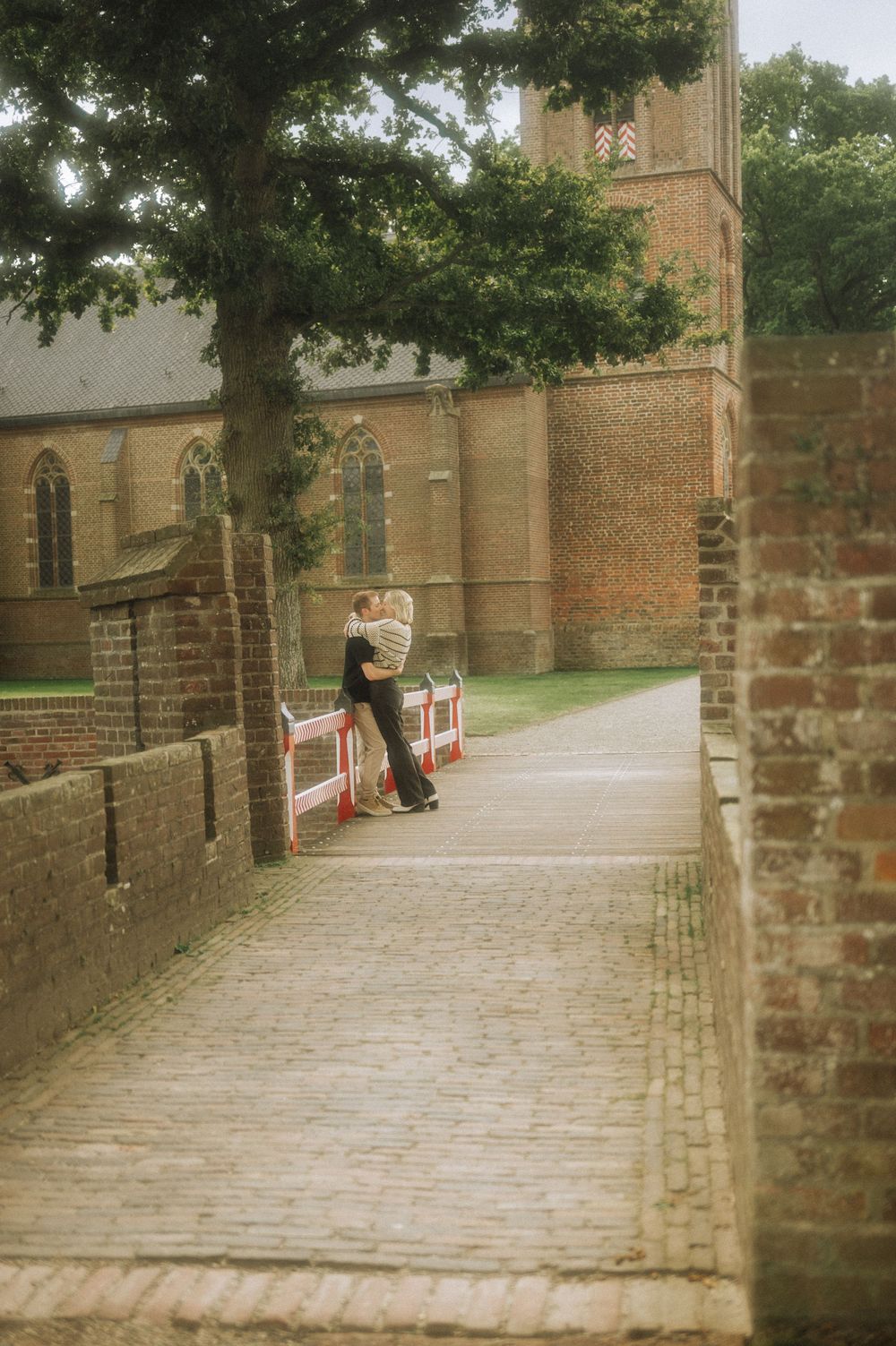 Brick-walled pathway with stone pillars leads to a historic building entrance while a person sits on a bench.