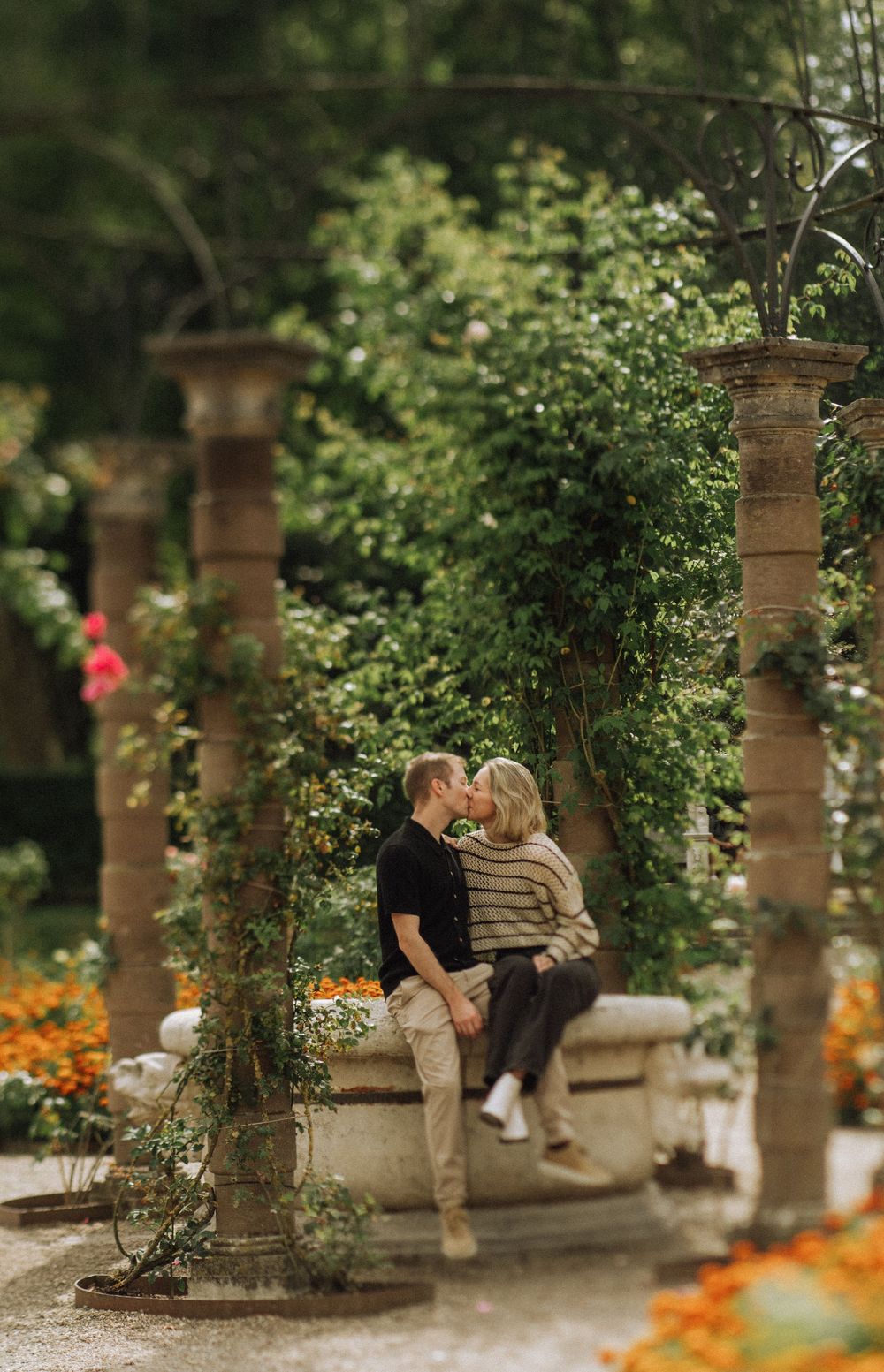 A romantic couple sits and kisses on a garden wall with stone pillars and orange flowers blooming below.
