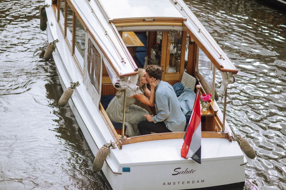 A wooden canal boat with red and white trim cruises through Amsterdam's waterways on a cloudy day.