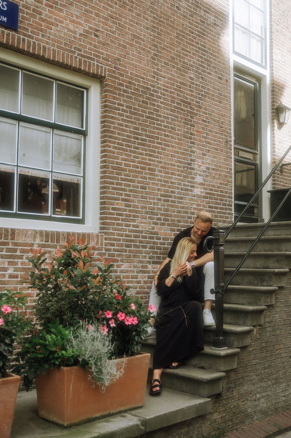 A person sits on outdoor brick steps near terracotta planters filled with pink flowers and greenery.