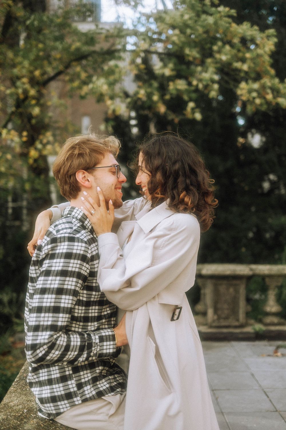 A couple shares a tender moment outdoors, one wearing a plaid shirt and the other in a white coat.