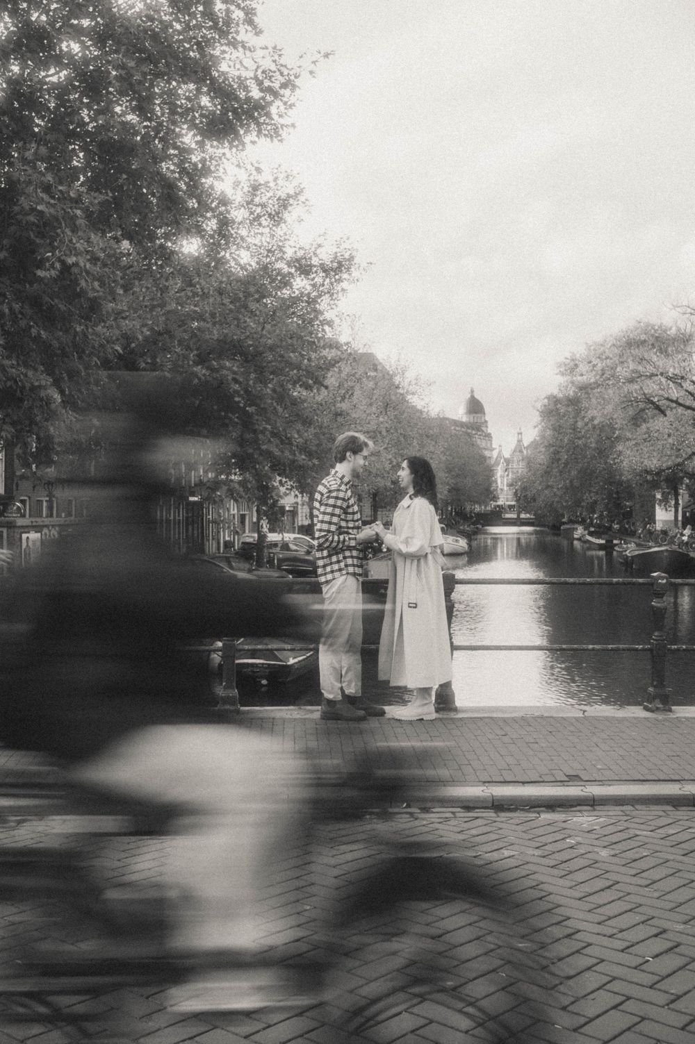 Black and white sequence showing motion blur of figures walking along a tree-lined cobblestone path near a canal.