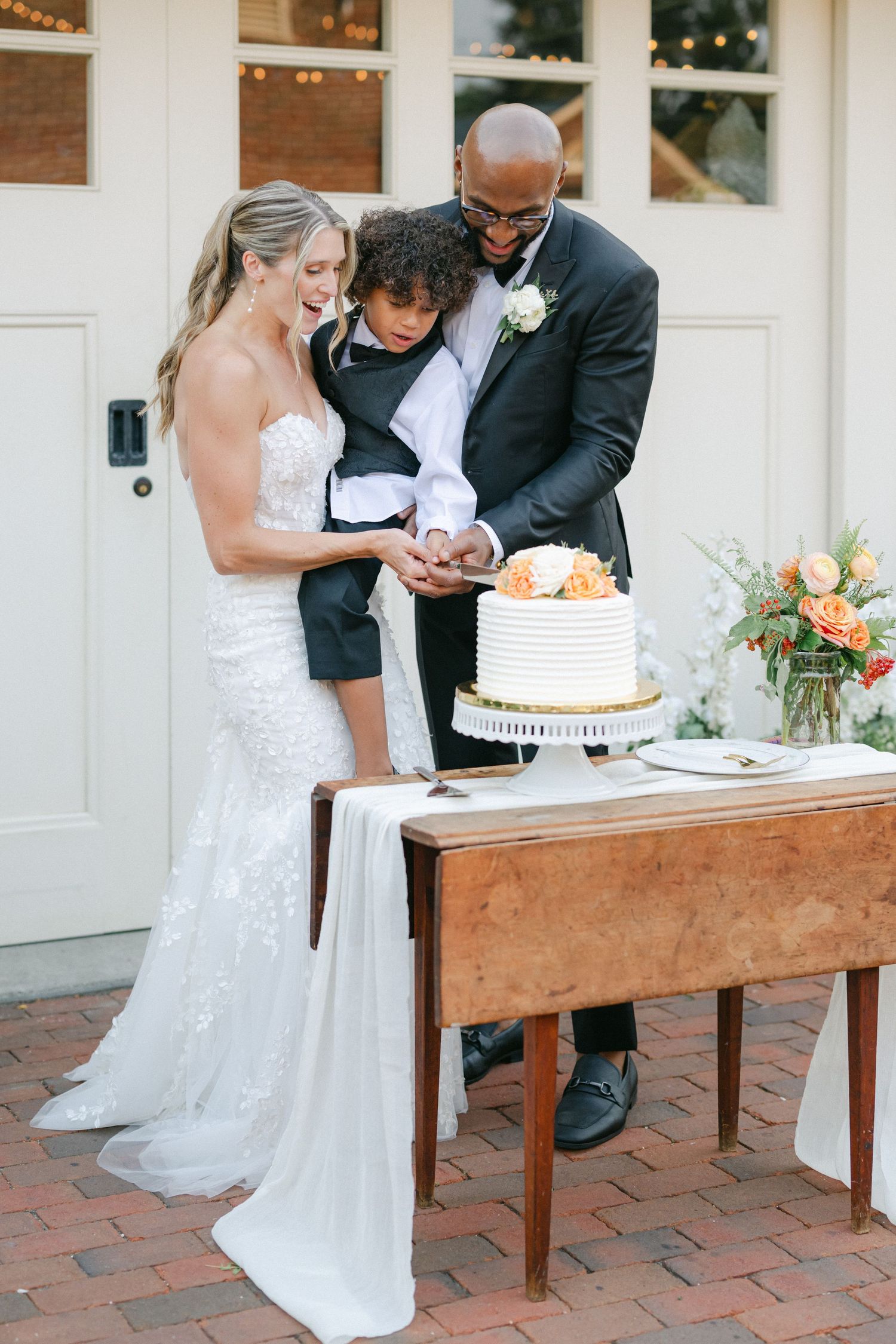 Wedding couple cuts their white tiered cake together at rustic wooden table on brick patio during outdoor reception. - Elawa Farm