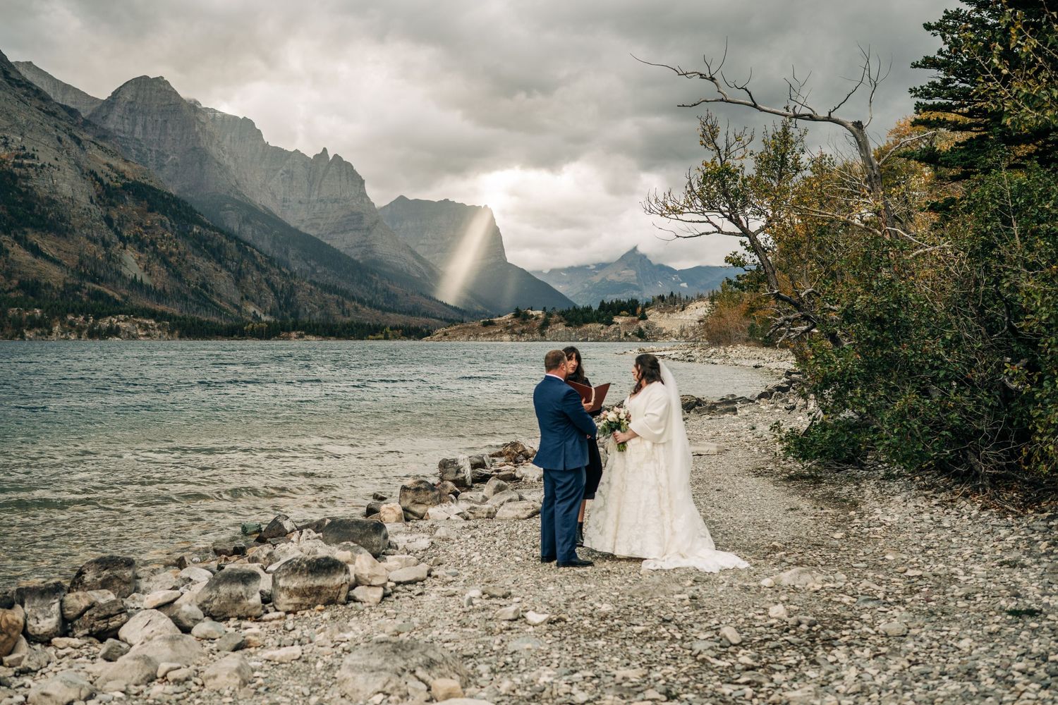 A mountain lake wedding ceremony on rising sun picnic area shoreline with dramatic cloudy skies and rocky shoreline in the background.