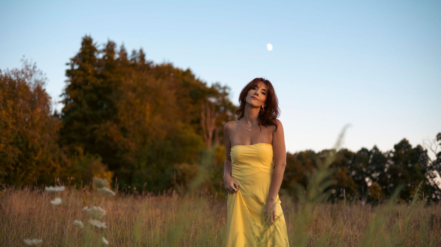 A woman in a yellow dress stands in a field of tall grass during golden hour with autumn trees in the background.