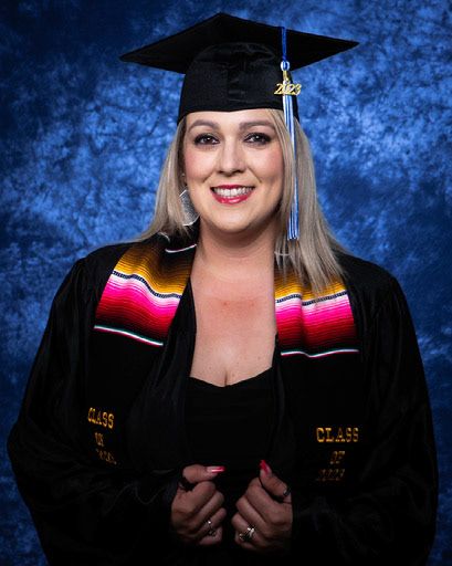 Graduate in black cap and gown poses against blue backdrop in professional graduation portrait series.