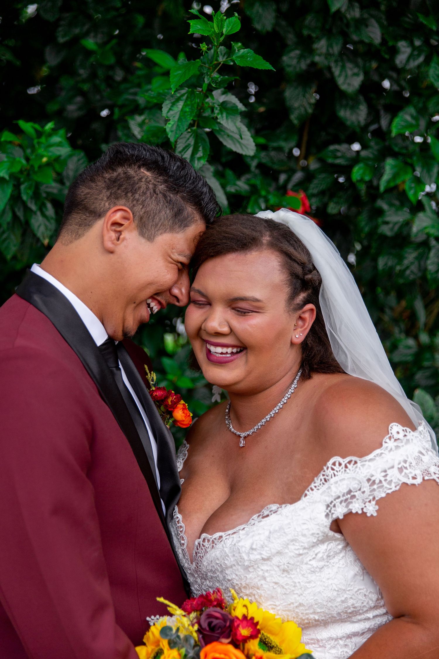 A joyful couple shares a candid moment of laughter on their wedding day, with colorful flowers and greenery at Heeia Park
