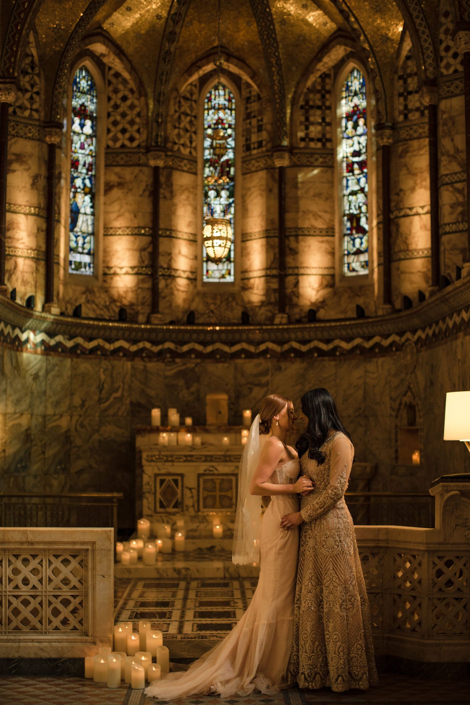 Two brides stand together in a stunning gothic cathedral with stained glass windows and glowing candlelight in Halifax Nova Scotia