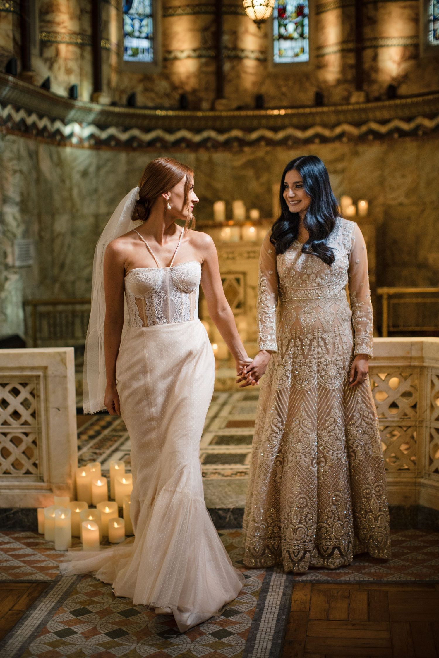 Two LGBTQ brides in elegant wedding gowns hold hands in a candlelit cathedral with stone columns and ornate details at Halifax wedding.