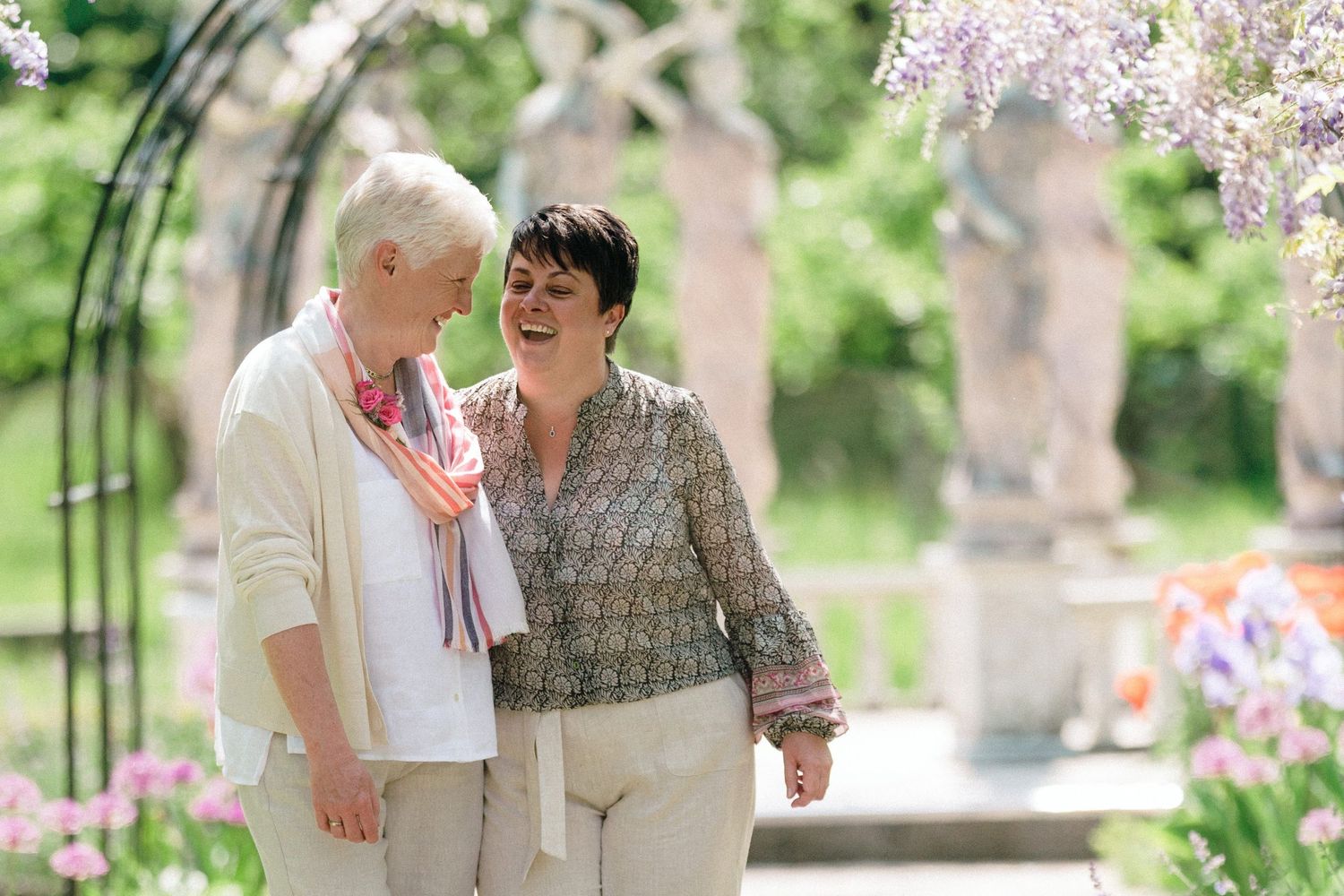 Two brides sharing a happy moment while walking together in a beautiful garden with blooming flowers and an arbor near The Lord Nelson Hotel