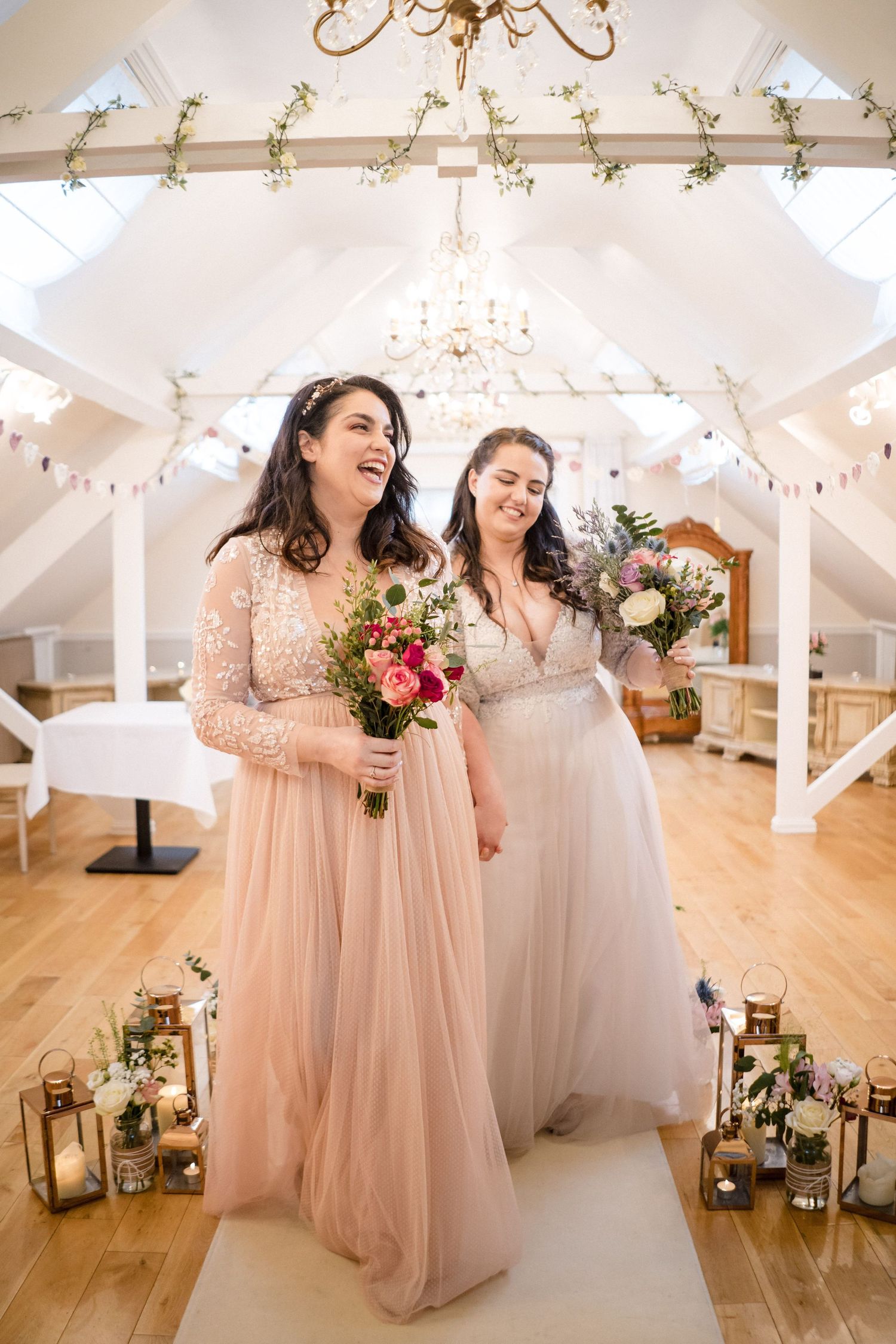 Two brides in flowing pink and white gowns share a joyful moment under draped white decor near The Lord Nelson, Halifax wedding venue.