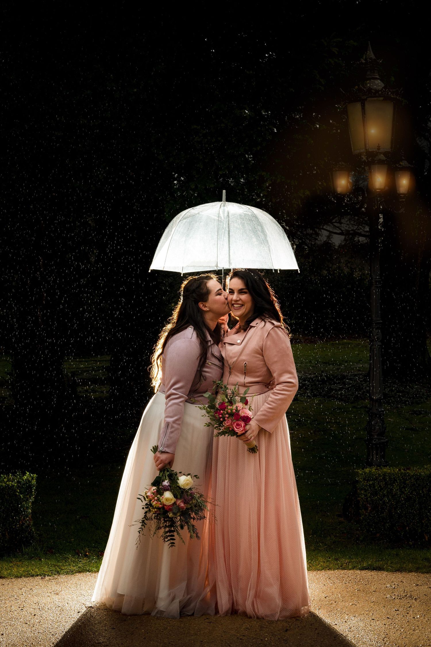 Two LGBTQ brides in pink dresses share an intimate moment under a white umbrella at night by glowing lanterns near The Lord Nelson, Halifax