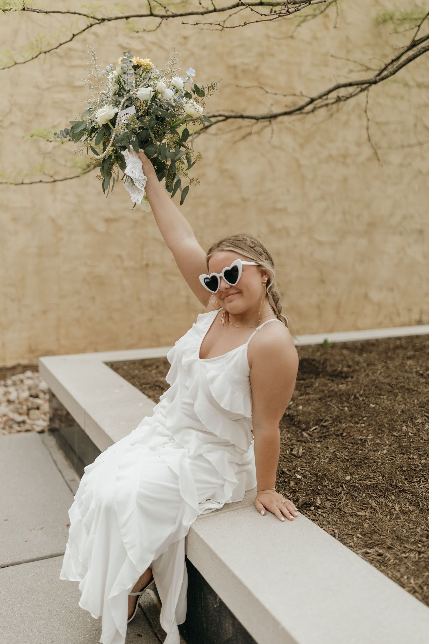 A person in a white dress and heart-shaped sunglasses holds up a bouquet while sitting on a concrete ledge.
