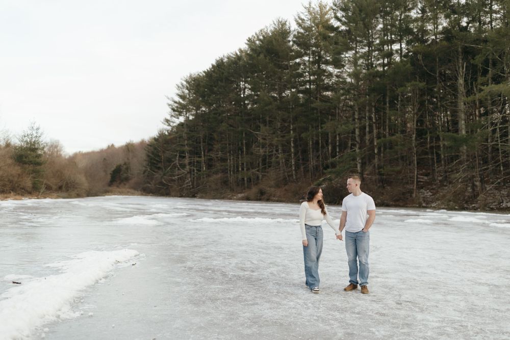 Couple standing together on frozen lake surface with pine forest backdrop.