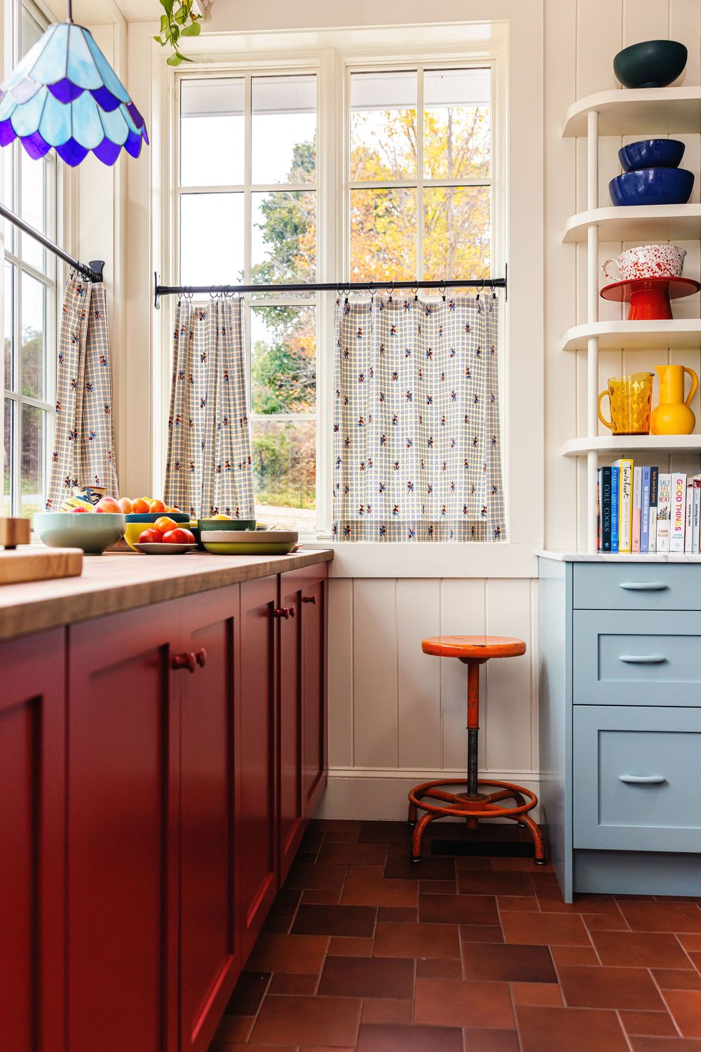 Interior design image of a colorful kitchen with red cabinets, blue drawers, open shelving, and a stained glass pendant light.