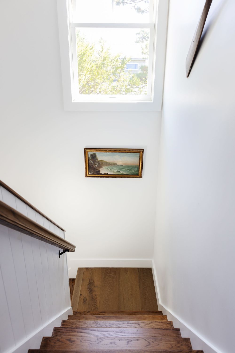 Wooden staircase with white walls, skylight window, and small landscape painting or an AirBnb listing on Fire Island.