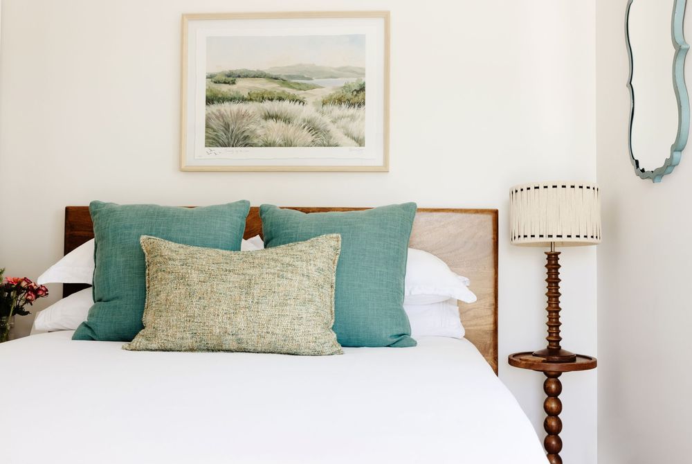 Bed with teal and green pillows, wooden headboard, landscape artwork, and vintage lamp or an AirBnb listing on Fire Island.