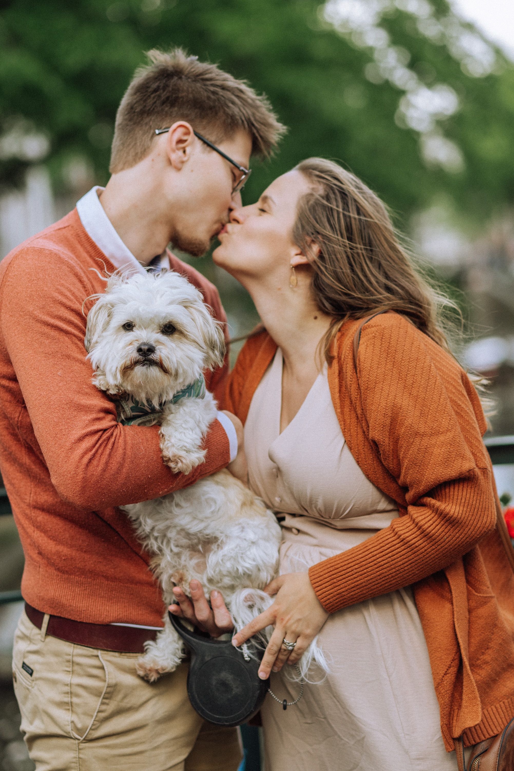 Outdoor maternity photos with couple in autumn sweaters holding small white dog among green foliage.
