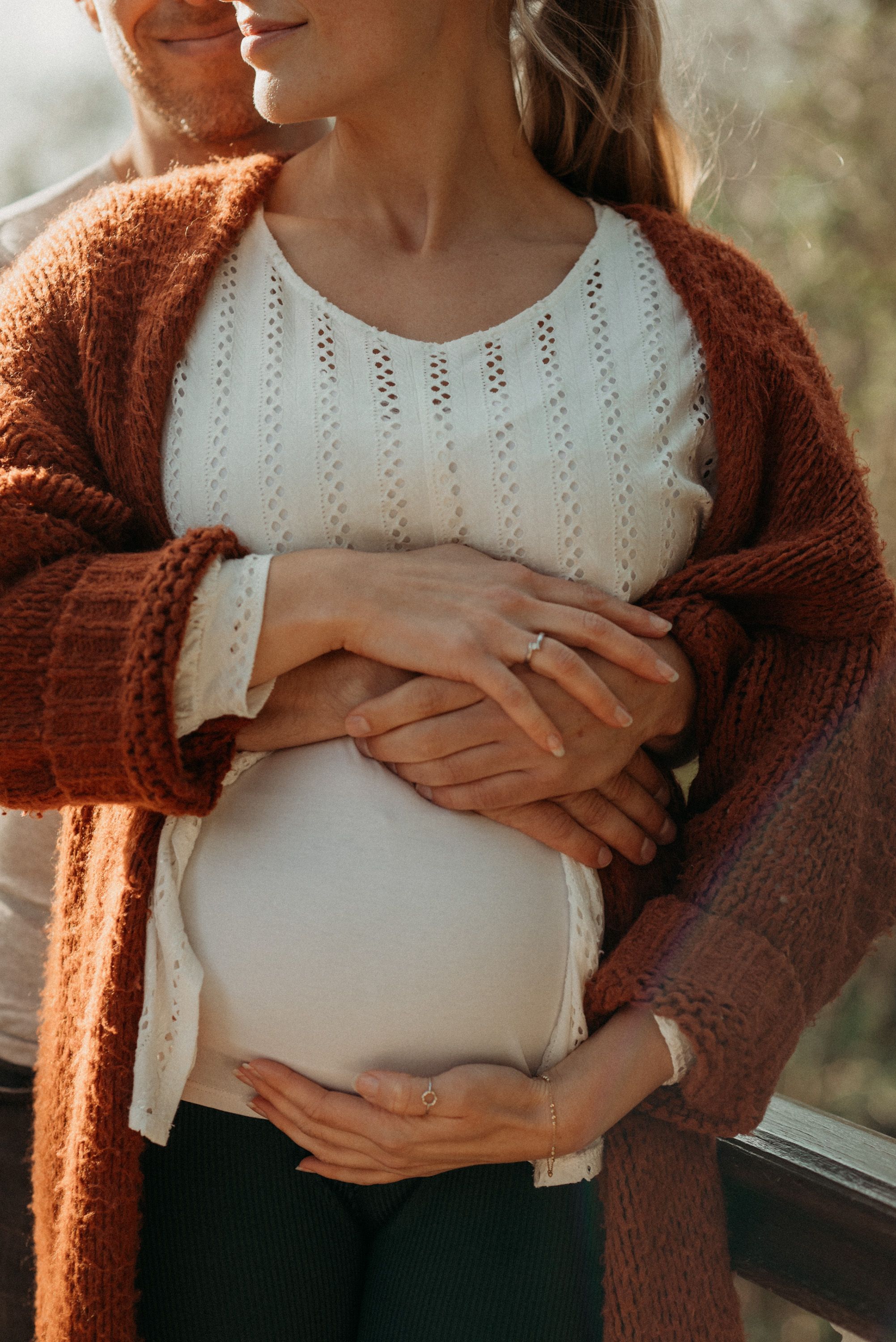 Close-up of hands resting on a pregnant belly wearing a white top and rust-colored cardigan.