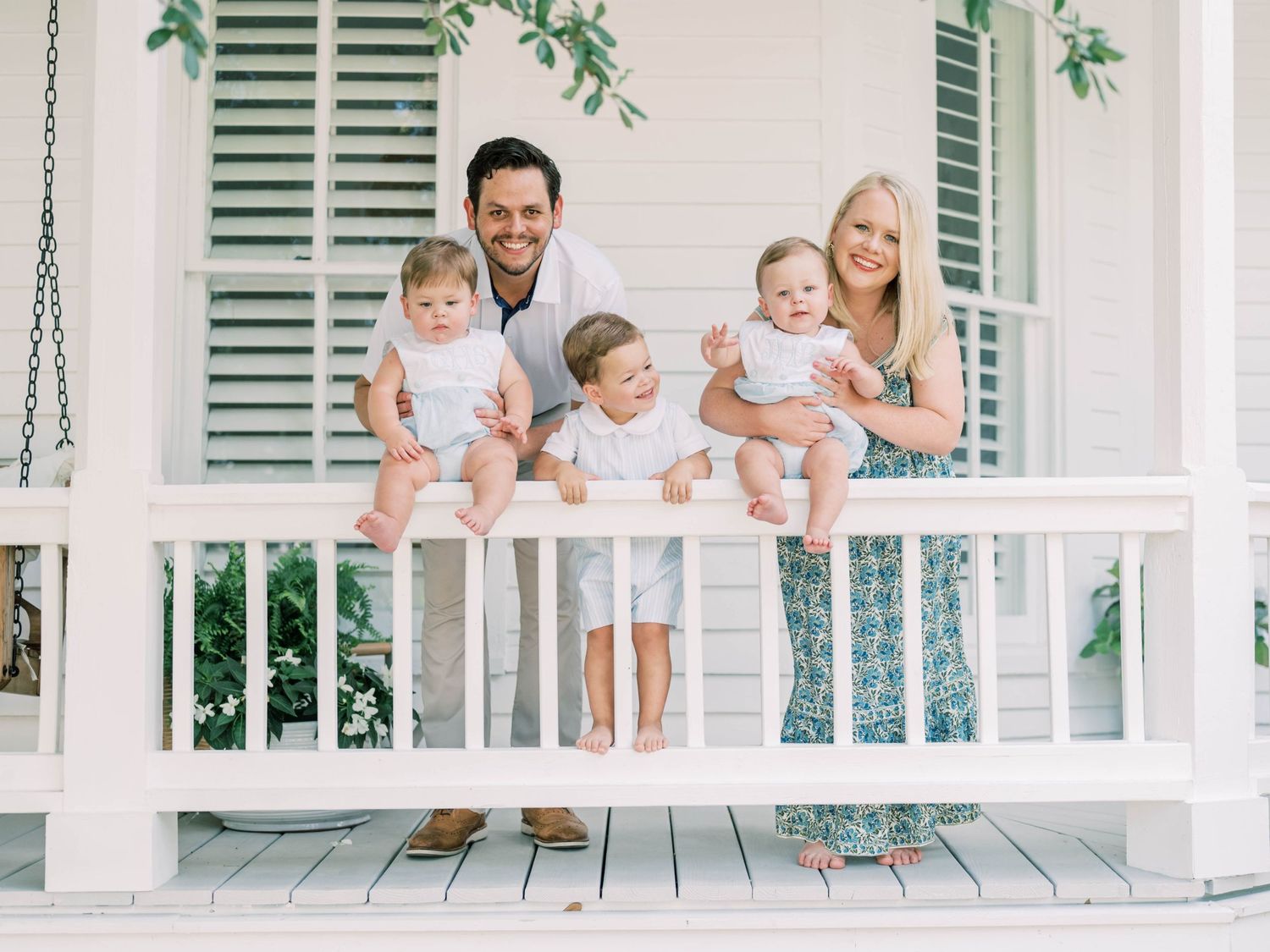 A family stands together on a white porch with traditional shutters and green vines growing above.