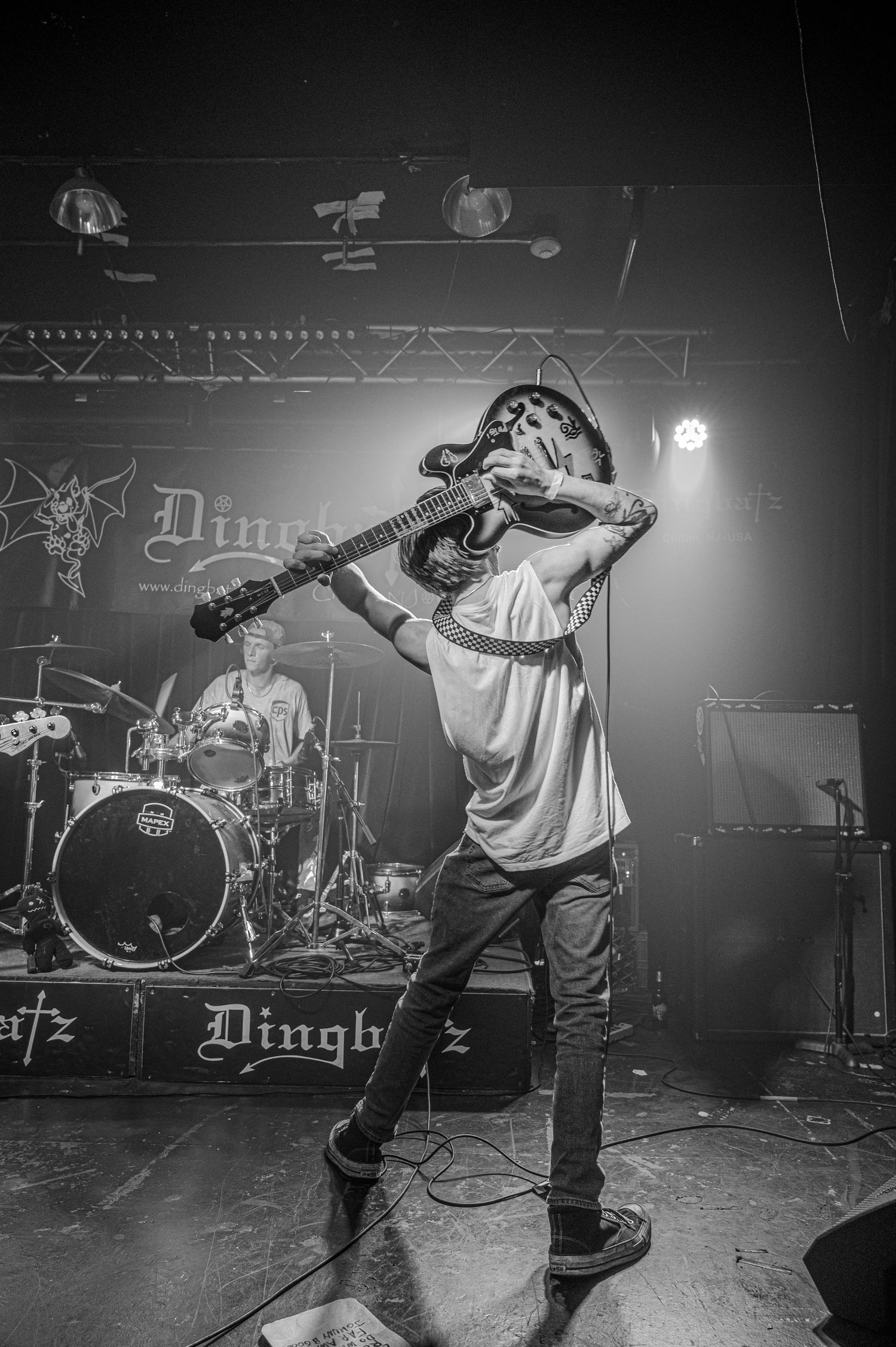 Black and white concert photo showing an energetic performer on stage with a guitar in front of drum kit and stage lighting.
