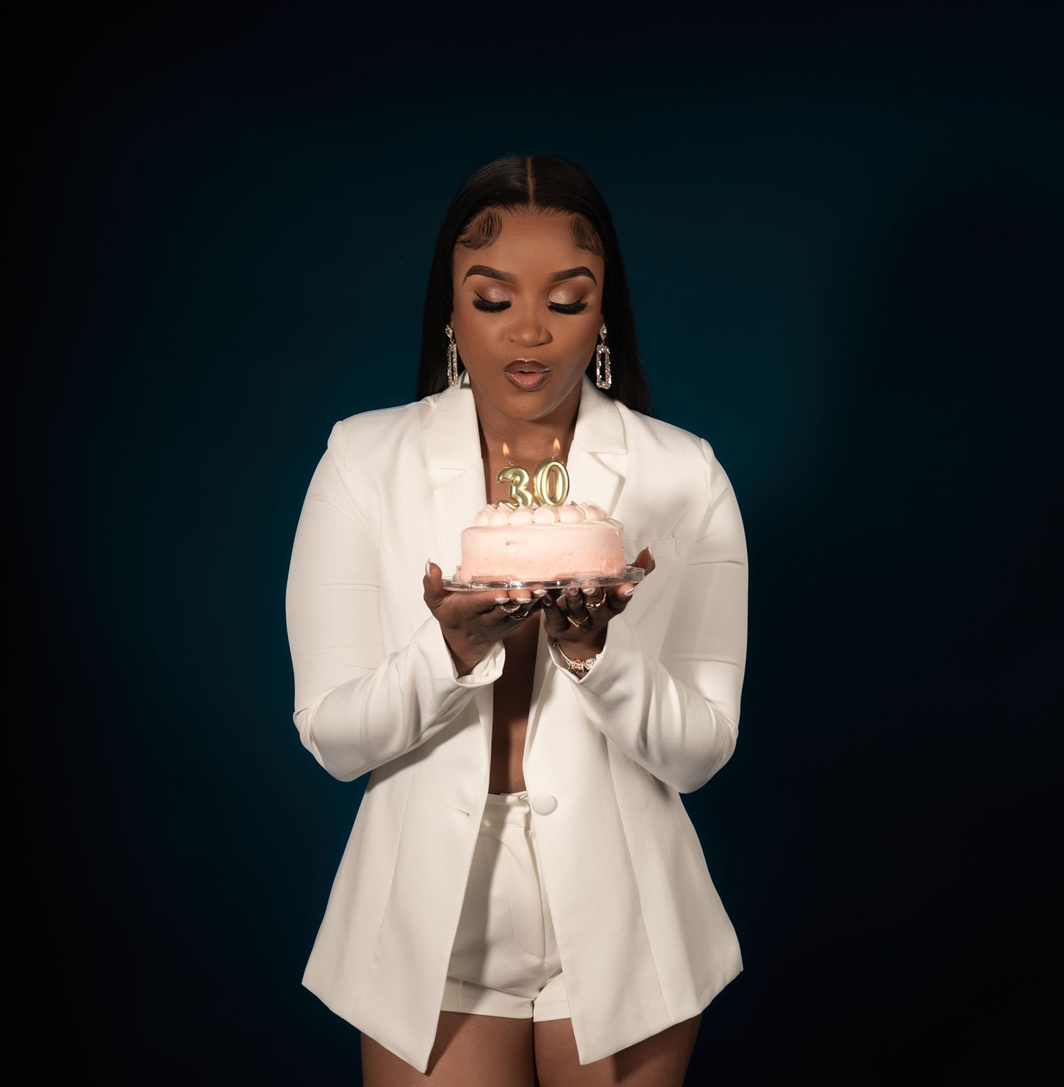 Person in white blazer outfit holds glowing birthday cake candles against dark background.