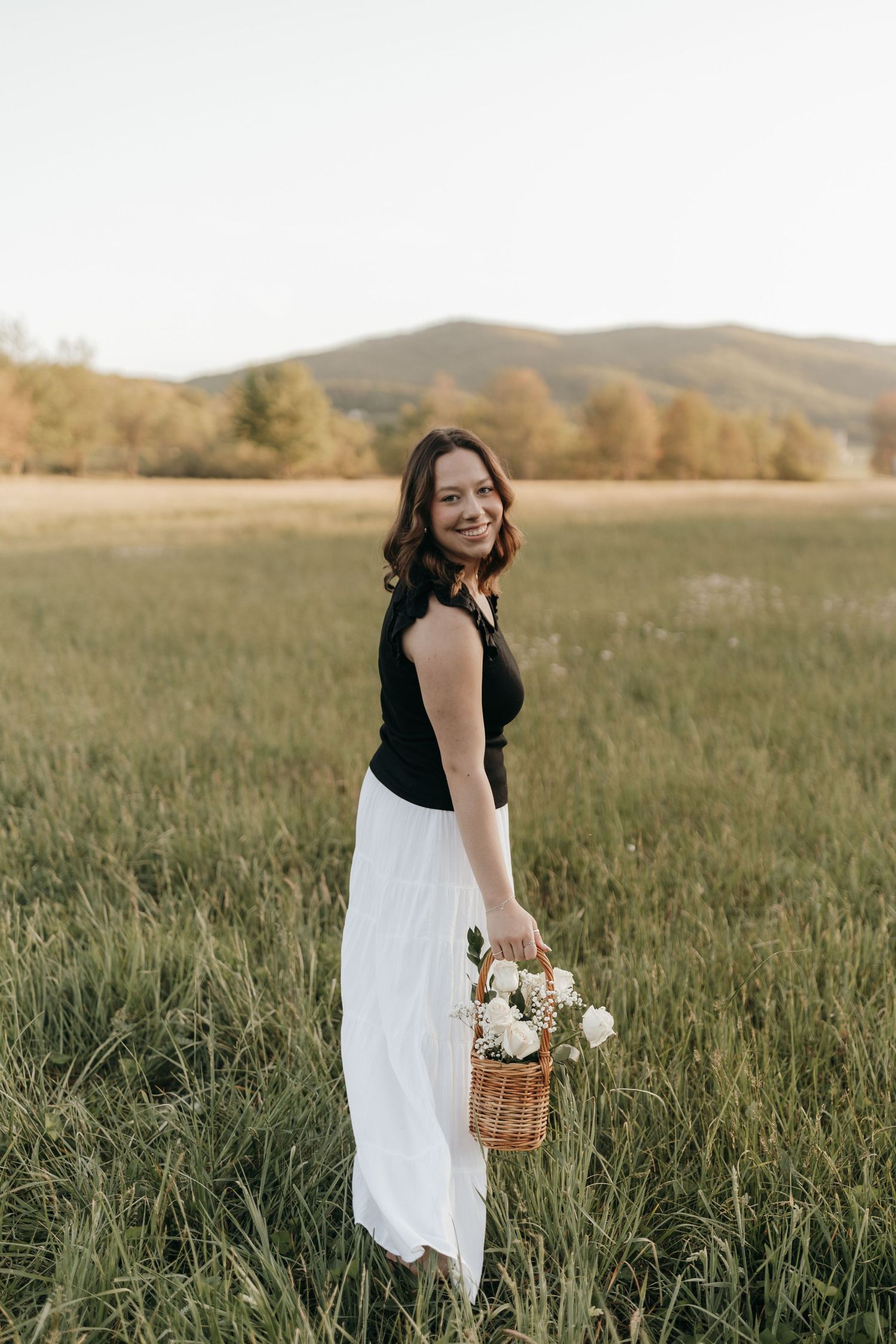 A woman in a black top and white skirt holds a basket of flowers while standing in a mountain field.  Virginia graduation/senior session.