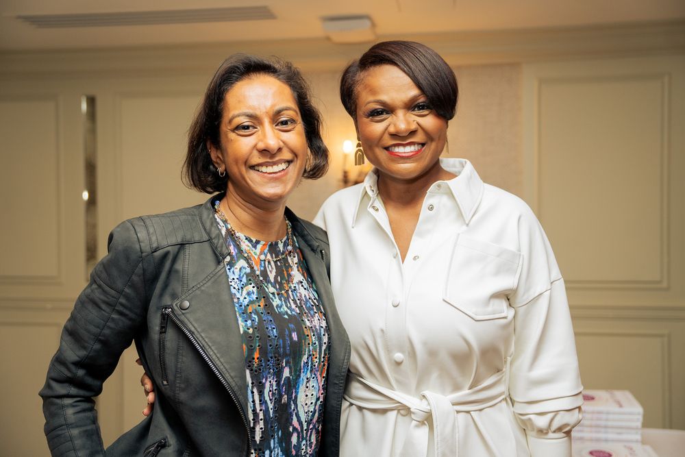 Two professional women smile warmly while standing together indoors wearing business attire.