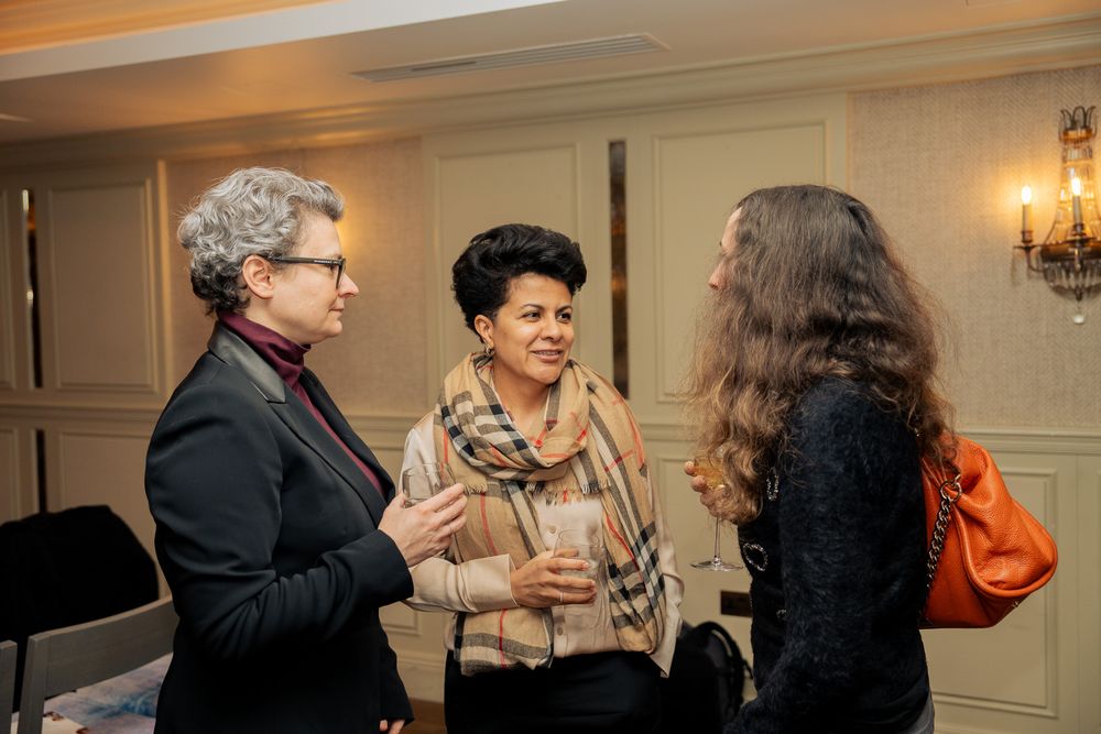 Three people engage in animated conversation at an indoor event with elegant wall sconces in the background.