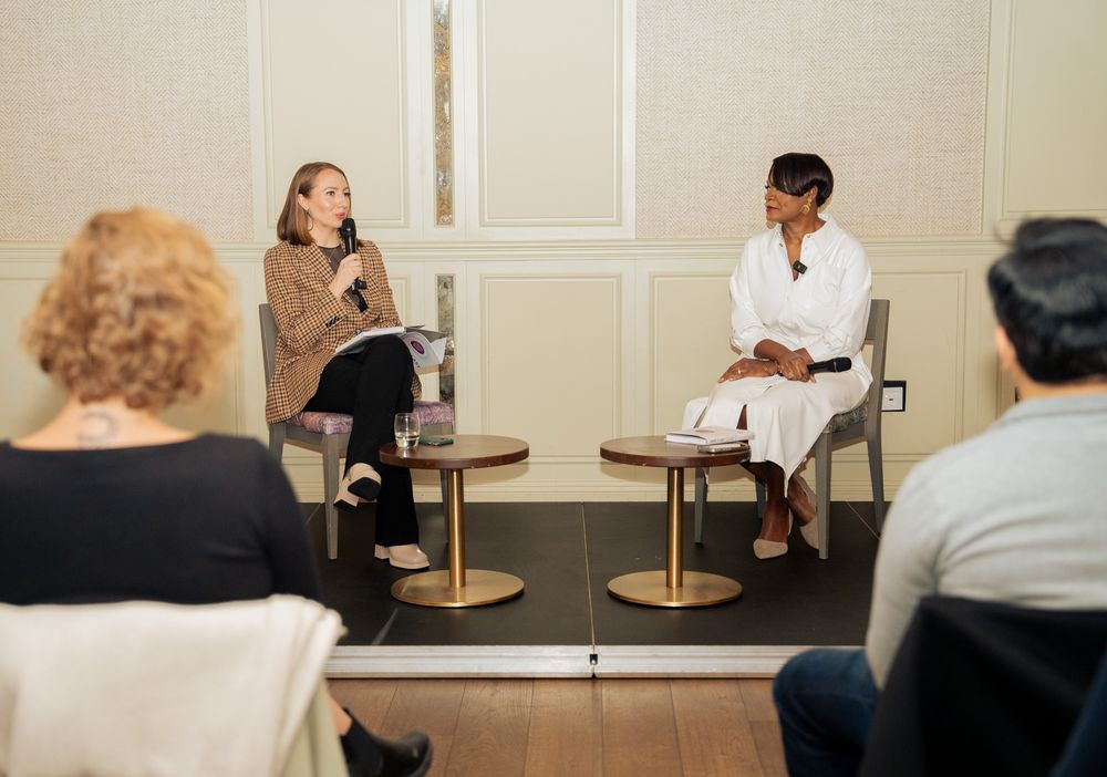 Two speakers sitting on stools having a discussion on stage while audience members watch in a bright room.