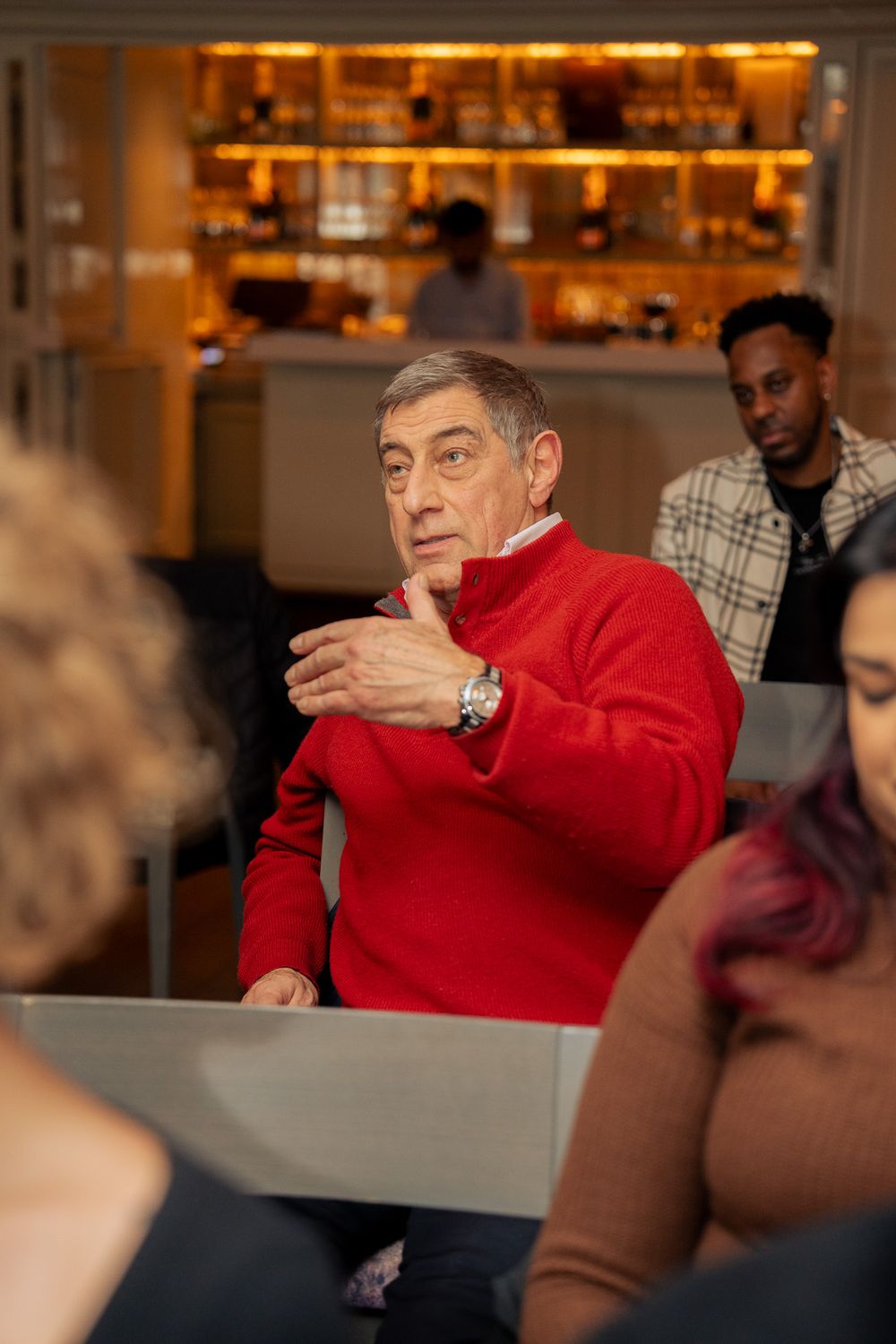 Person in bright red sweater gestures animatedly while speaking at a social gathering in a bar setting.
