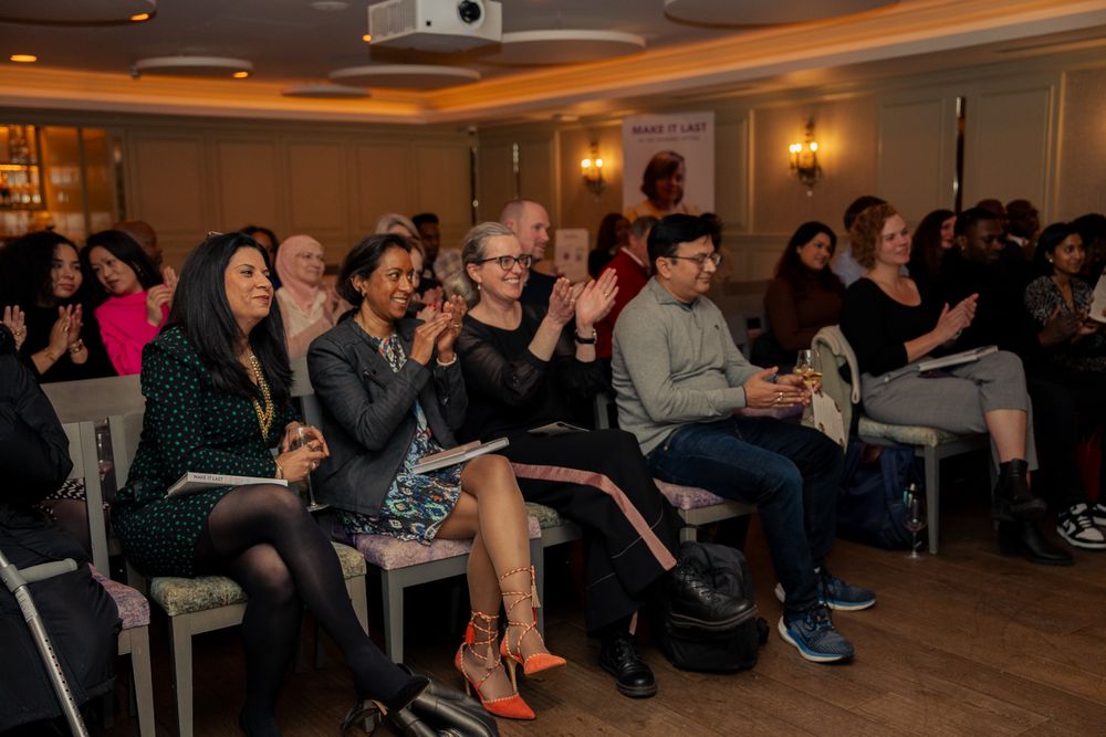 Audience members seated in rows watch and applaud during an indoor event in a warmly lit venue.