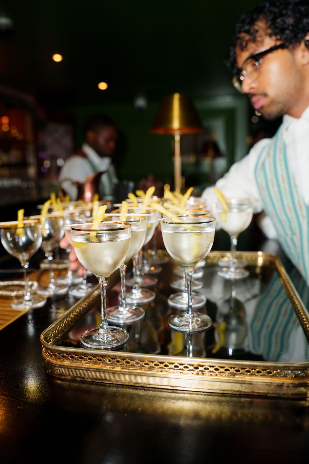 Bartender arranging elegant martini cocktails with garnishes on a gold tray at an upscale bar.
