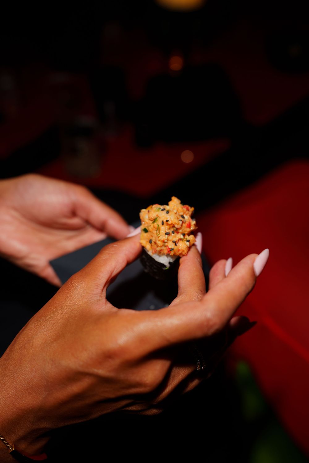 Hands holding a temaki sushi cone filled with spicy salmon and sesame seeds at a restaurant.
