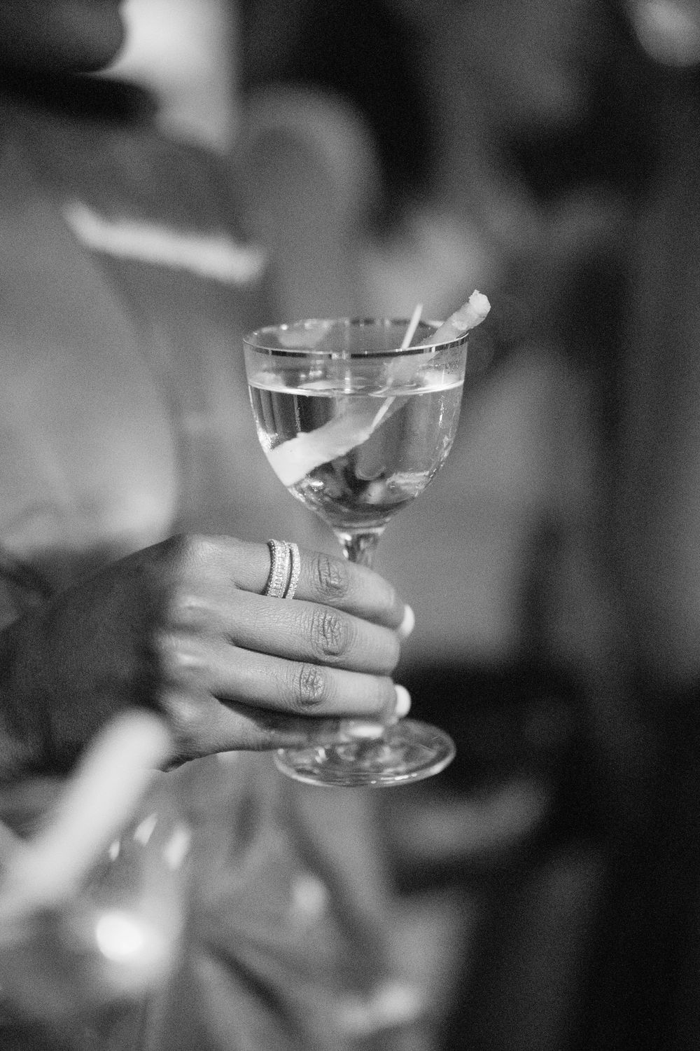 Black and white photo of a hand with rings holding an elegant cocktail glass with a citrus garnish.