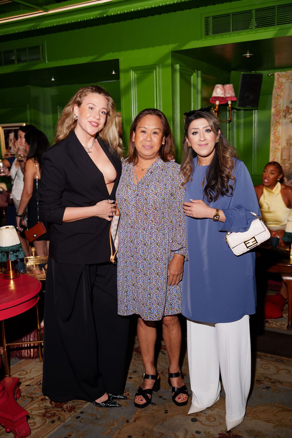 Three women posing together at an indoor event, dressed in black, floral, and blue outfits against a green wall.